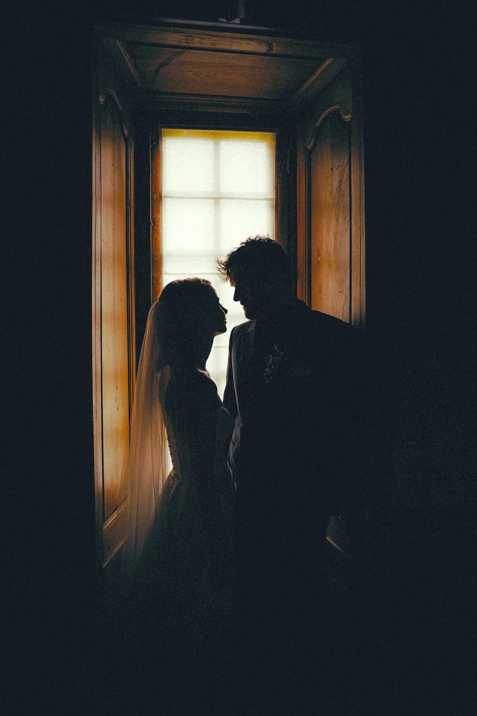Silhouettes d'un couple en gros plan, devant une fenêtre avec rideau lumineux, dans une pièce sombre avec murs en bois.