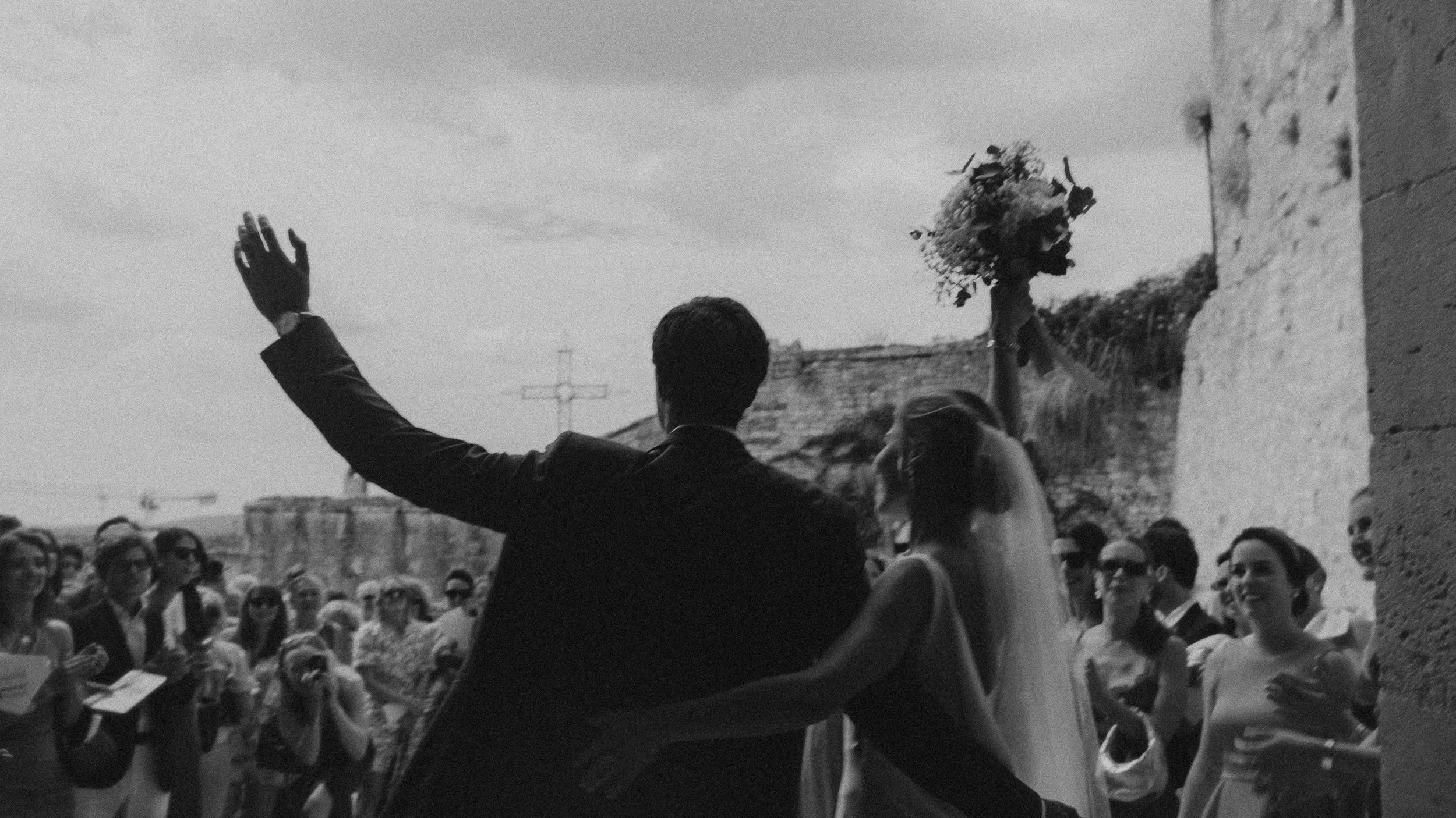 Un homme en costume noir levant la main devant une foule lors d'une cérémonie en plein air, avec une femme portant un bouquet, dans un environnement historique en pierre, en noir et blanc.