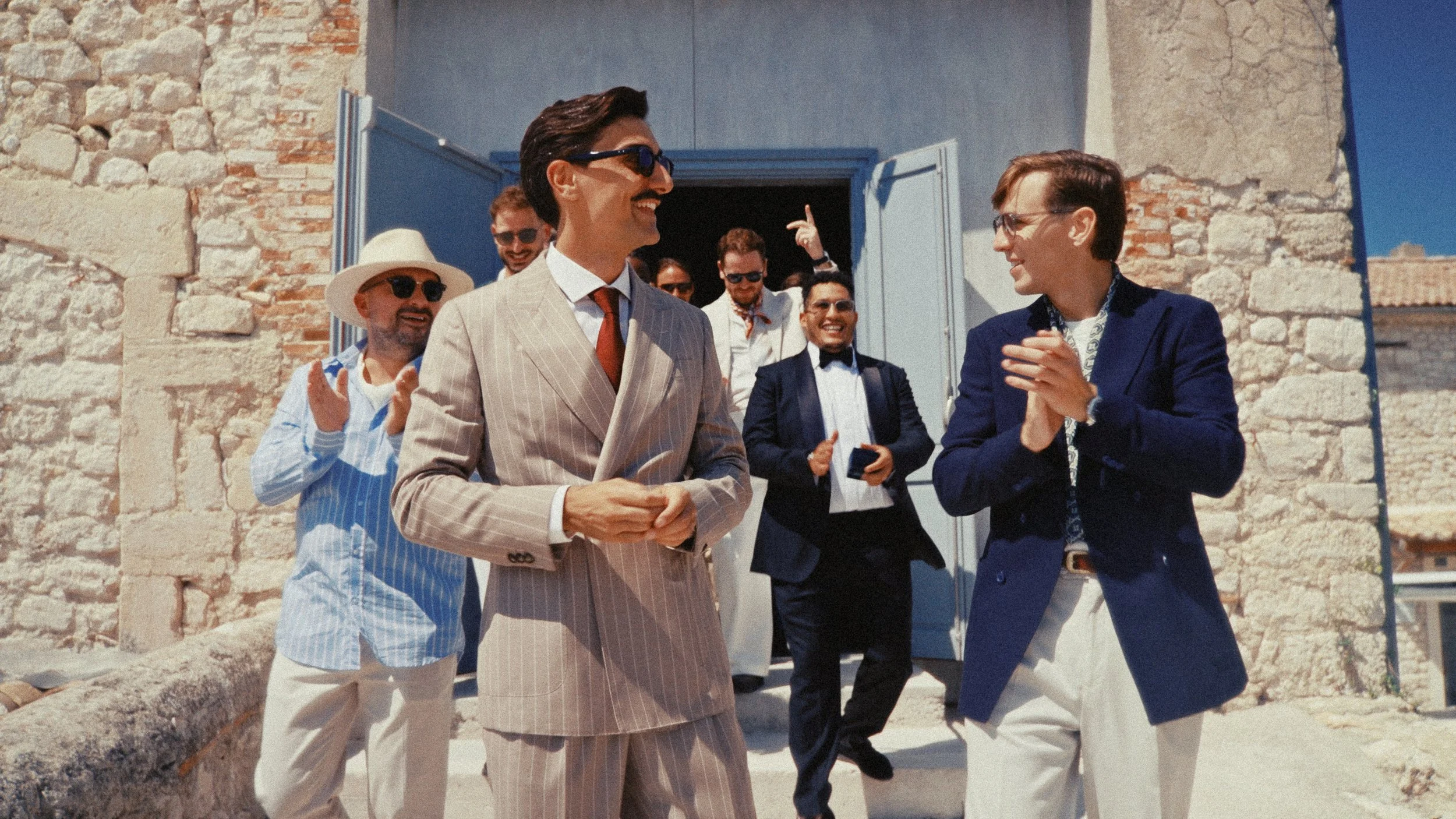 Groupe de personnes élégamment habillées, en costume et en robe, sortant d'un bâtiment en pierre sous un ciel bleu. Ils sourient, discutent et semblent heureux.
