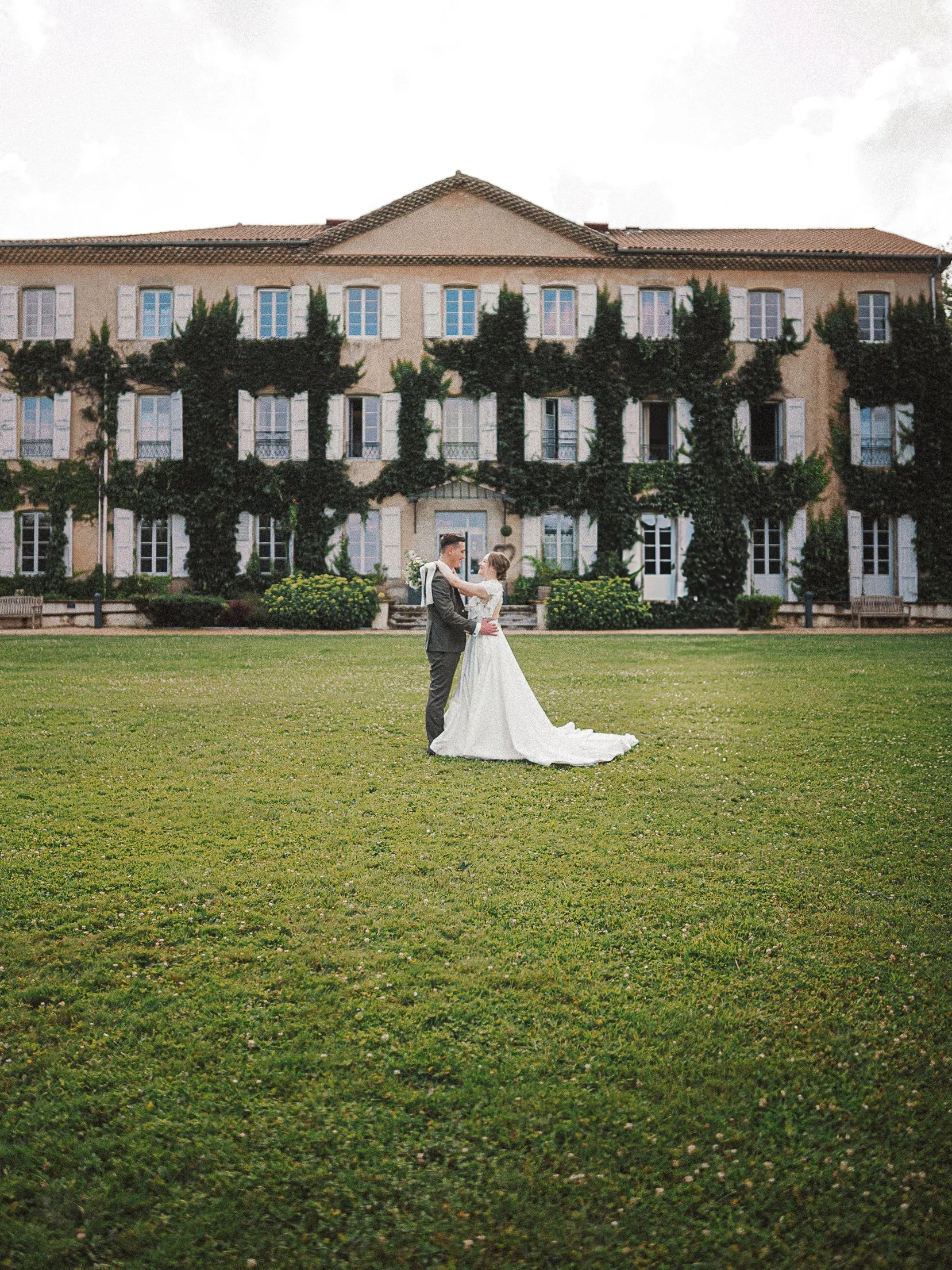 Un couple en robe de mariage danse sur une pelouse devant un grand bâtiment recouvert de vignes.