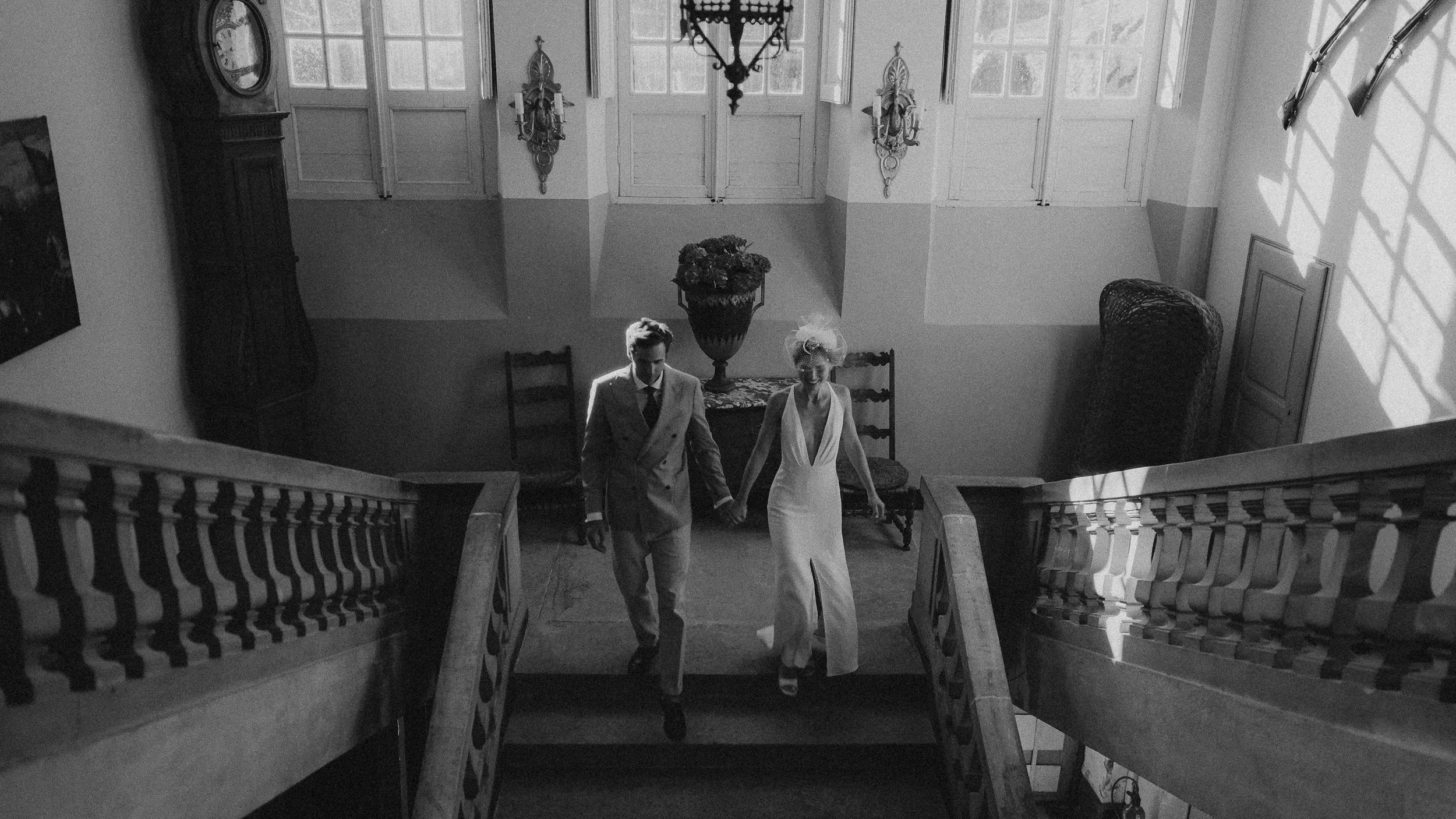 Deux personnes, un homme et une femme, marchent main dans la main dans un intérieur ancien, avec un escalier en bois, une grande fenêtre, et des lampes anciennes sur le mur, en noir et blanc.