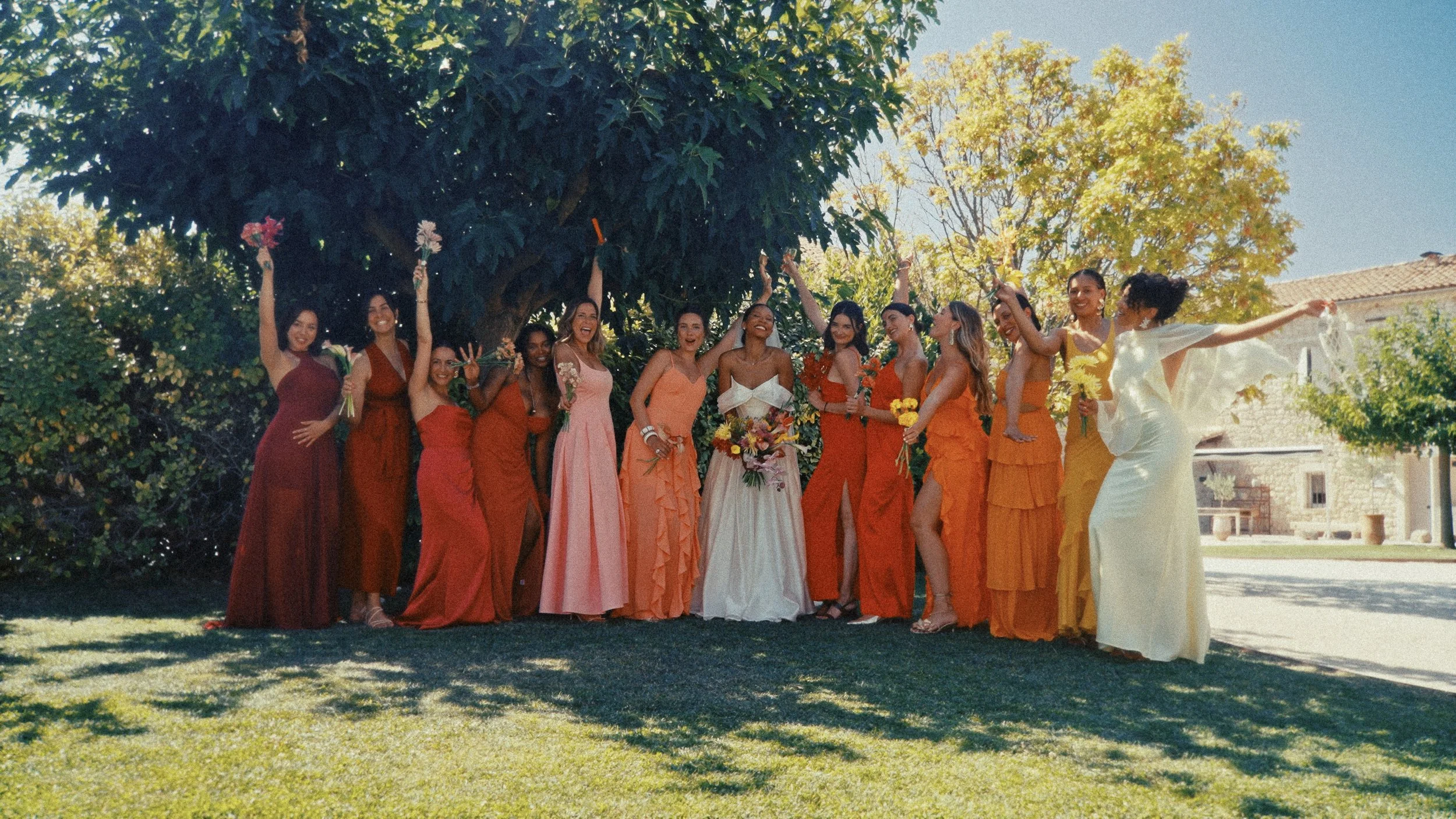 Un groupe de femmes en robes colorées posant sous un arbre lors d'une célébration festive en plein air, avec un ciel ensoleillé et des bâtiments en pierre en arrière-plan.