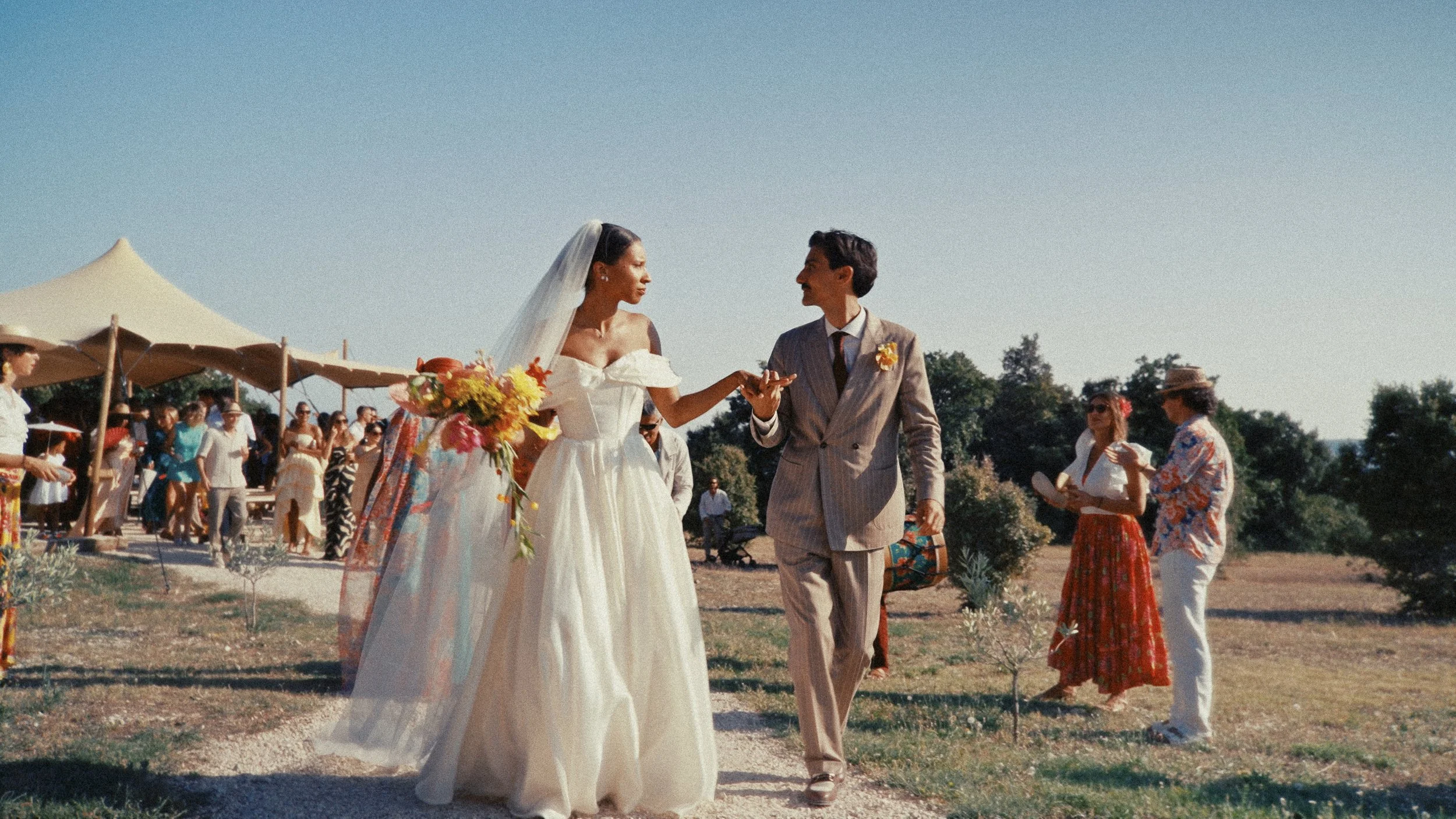 Un couple de mariés marchant main dans la main lors d'une cérémonie en plein air, entourés de convives sous de grandes tentes, par une belle journée ensoleillée.