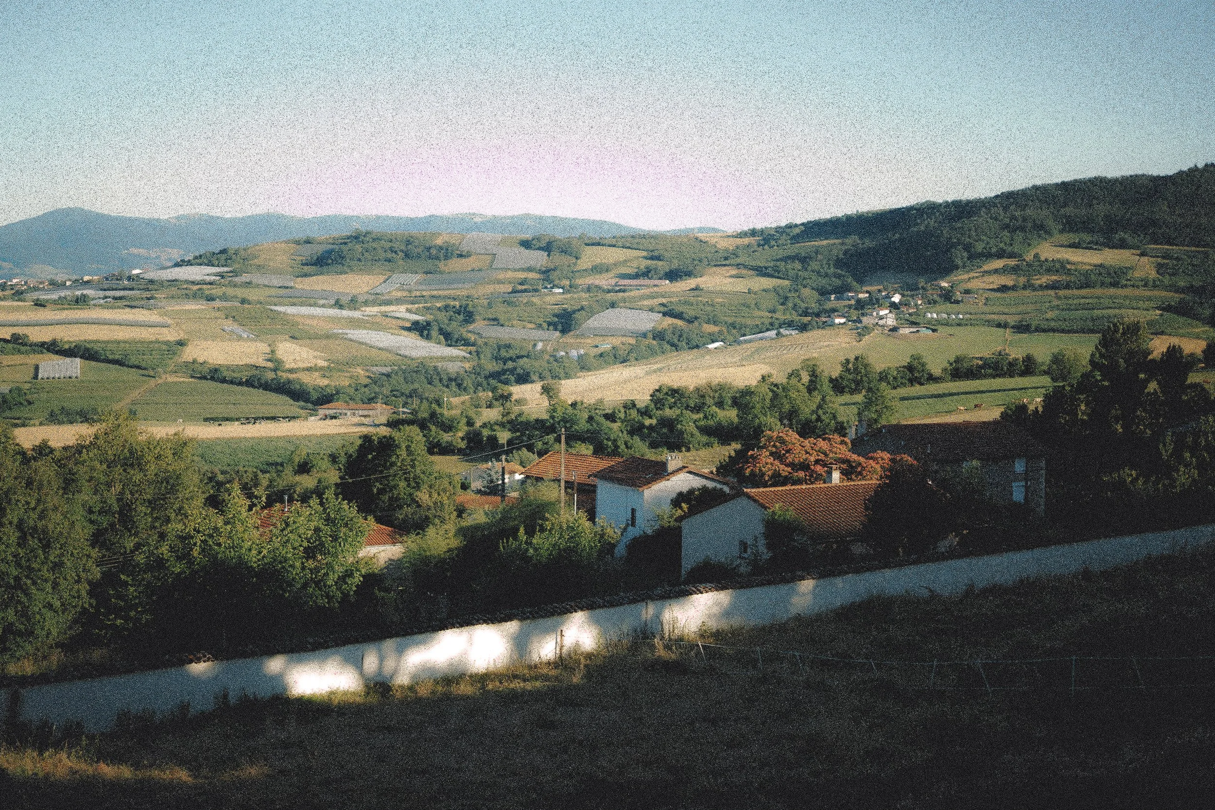 Paysage rural avec maisons, arbres et montagnes en arrière-plan sous un ciel clair.