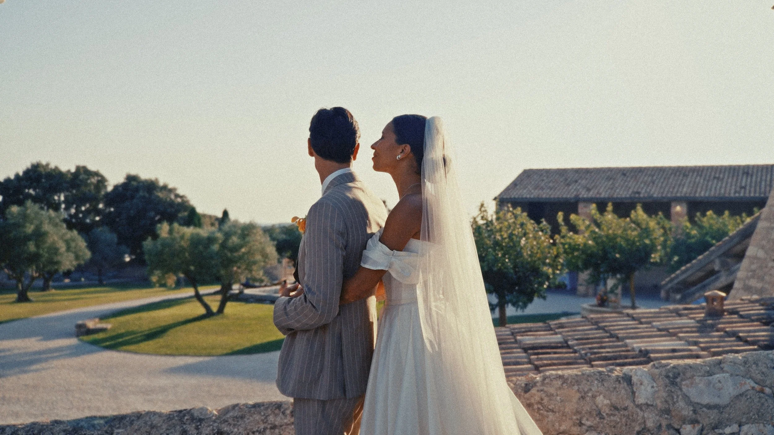 Un couple de mariés se tient près d'un mur en pierre, de dos, avec une vue sur un jardin et un bâtiment en arrière-plan, lors d’une cérémonie de mariage en plein air au coucher du soleil.
