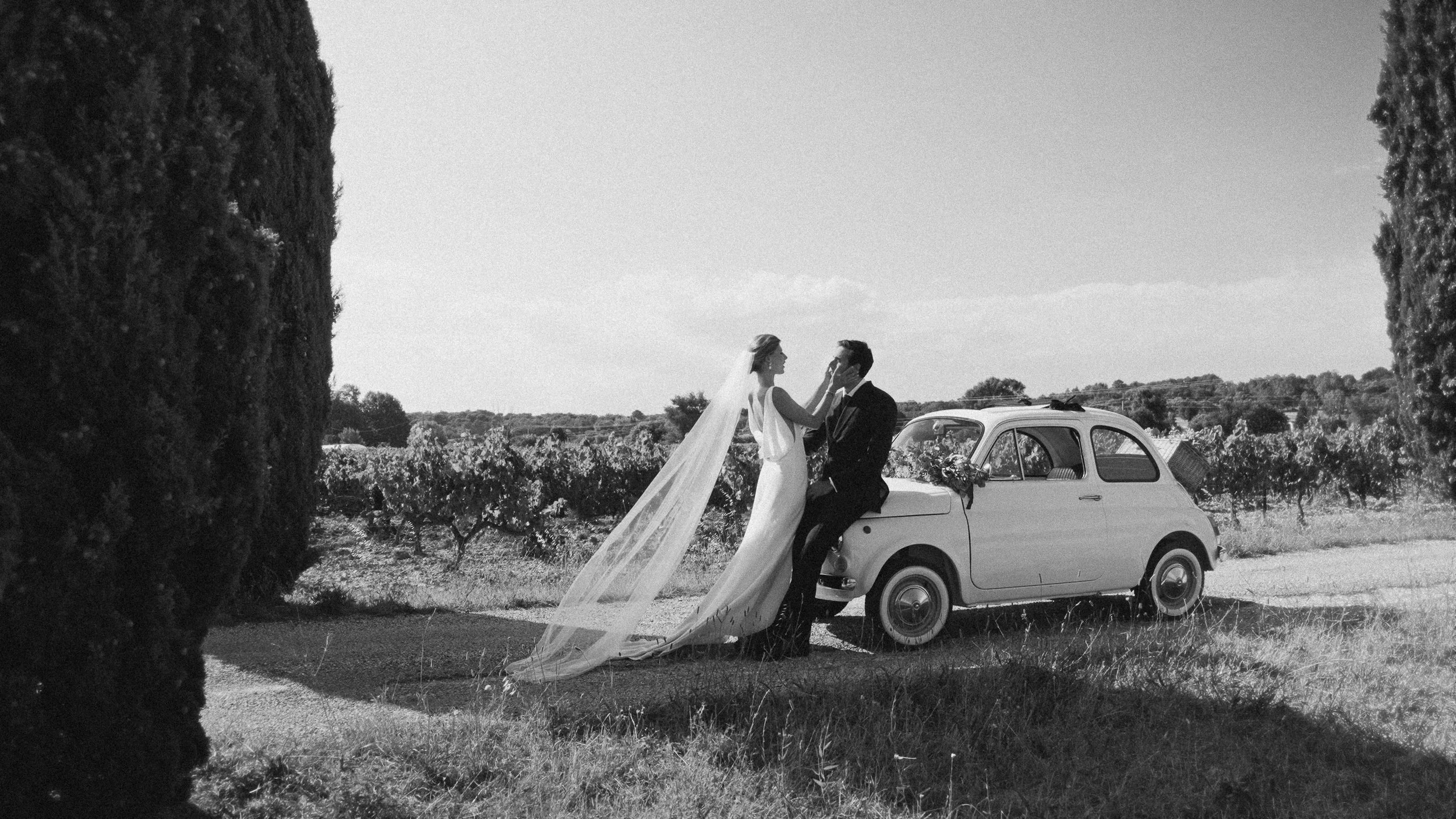 Une mariée et un marié en tenue de mariage devant une voiture vintage dans un paysage rural, en noir et blanc.