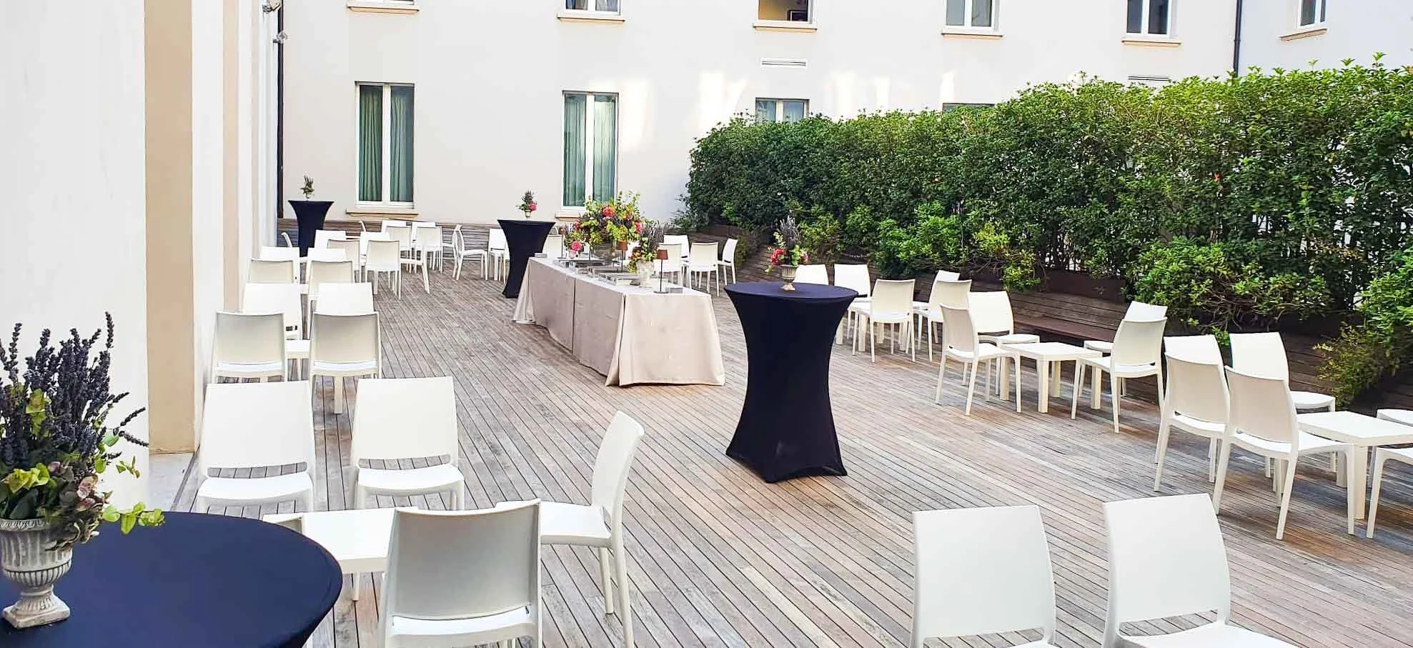Terrazzamento all'aperto allestito per un evento, con tavoli e sedie bianche e alte tavoli neri, decorati con fiori, e ampie siepi verdi sullo sfondo.