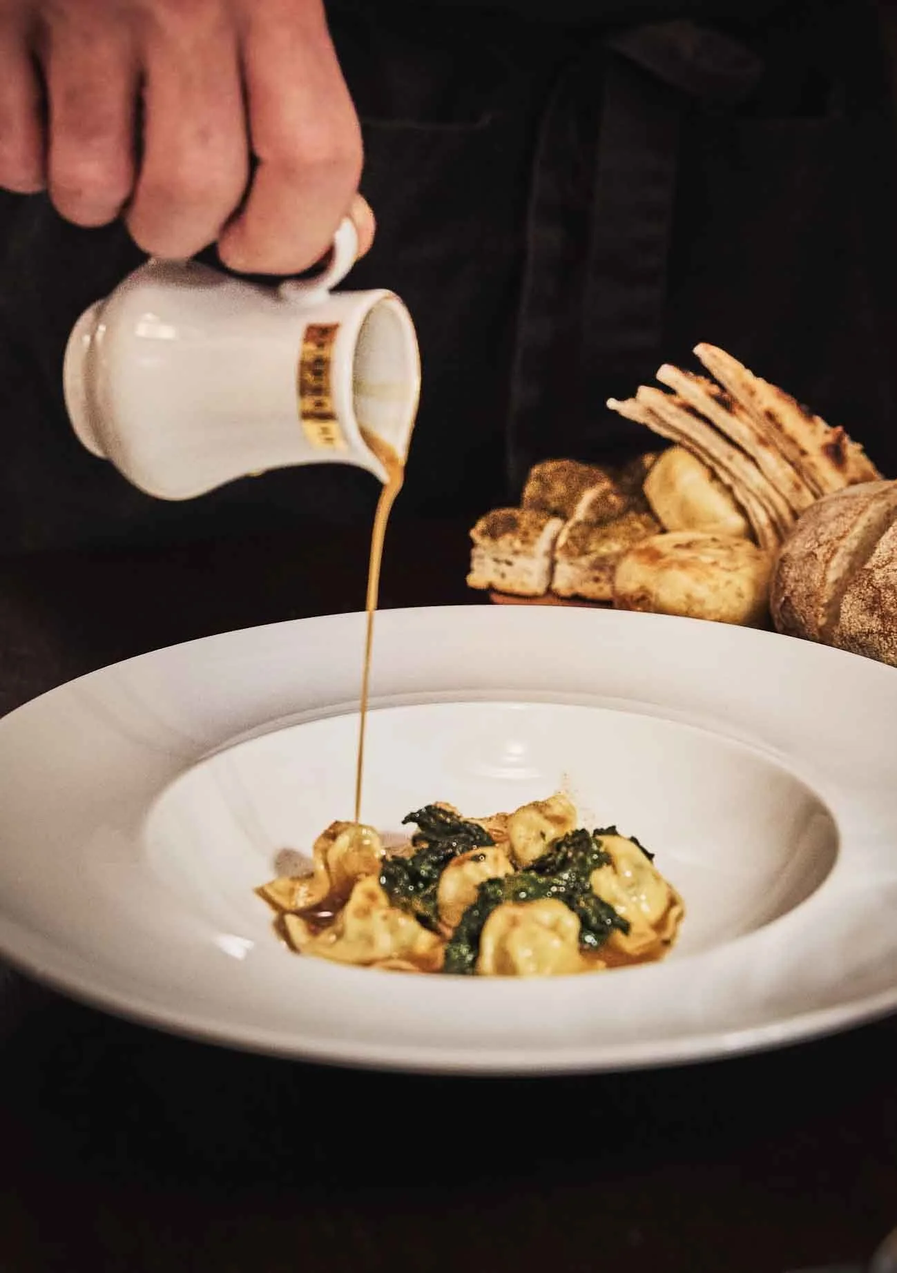 Un piatto di pasta con salsa verde, probabilmente con erbette e formaggi, decorato con salvia o altre erbe, con olio che viene versato sopra. Sullo sfondo, pane e pane rustico, con una mano che versando olio o salsa.