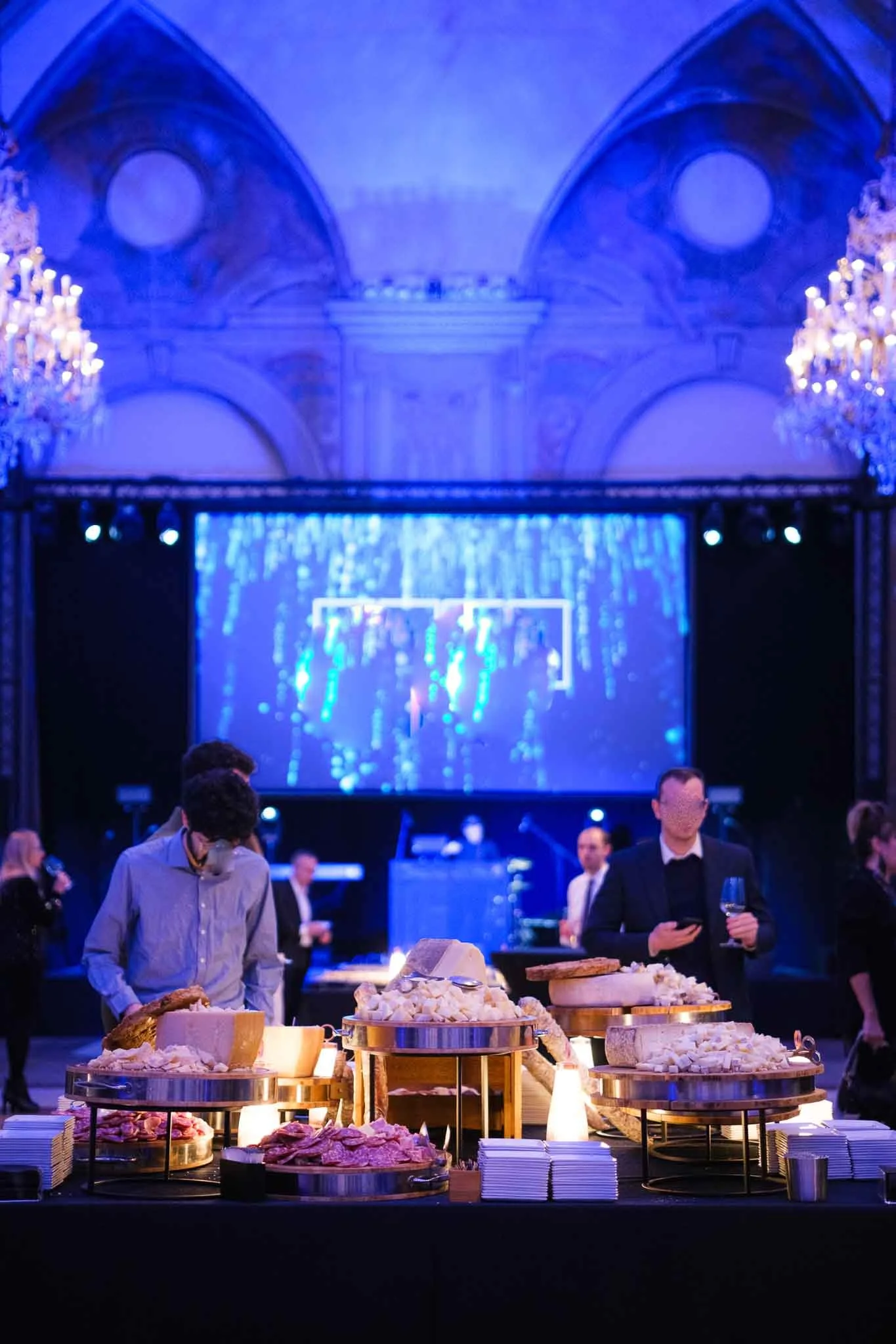 Una scena di un evento elegante con un buffet di formaggi e salumi, persone che conversano e un grande schermo al centro, decorazioni di luci e ghirlande.
