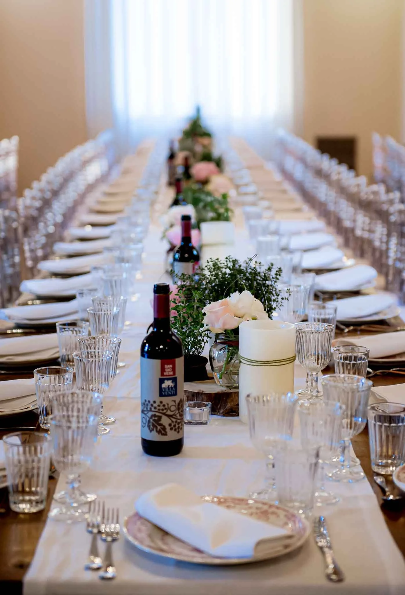 Tavolo apparecchiato per una festa o un evento, decorato con fiori, candele, bottiglie di vino e calici, con posti a sedere lungo entrambi i lati.