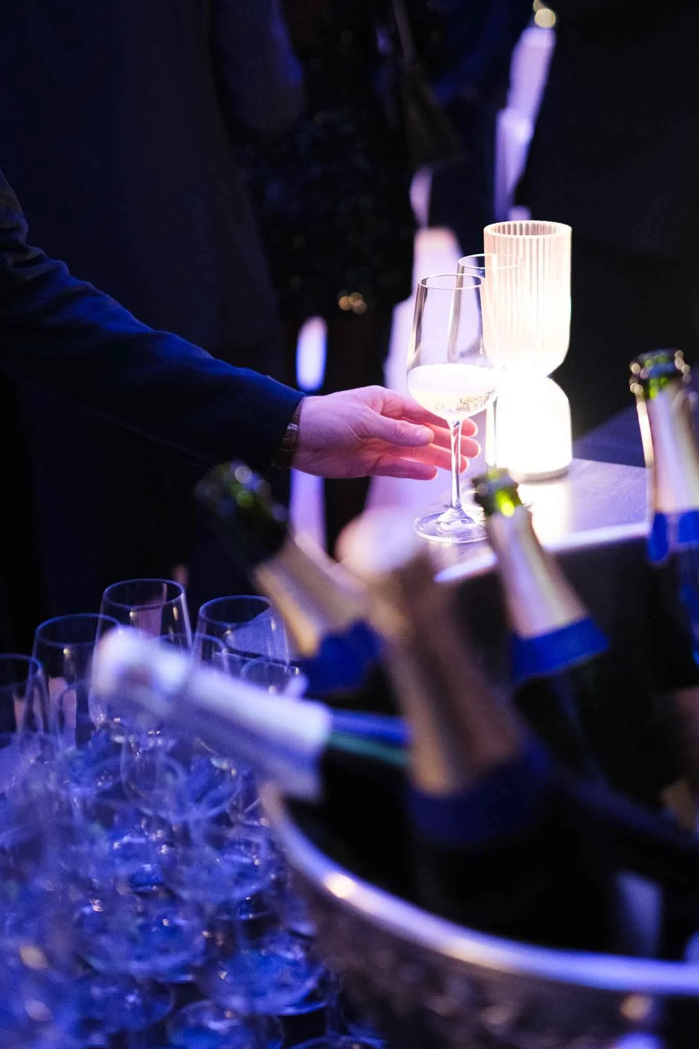 Persona che serve un bicchiere di vino bianco a tavola durante una festa o un evento notturno, con luci soffuse e illuminazione decorativa.