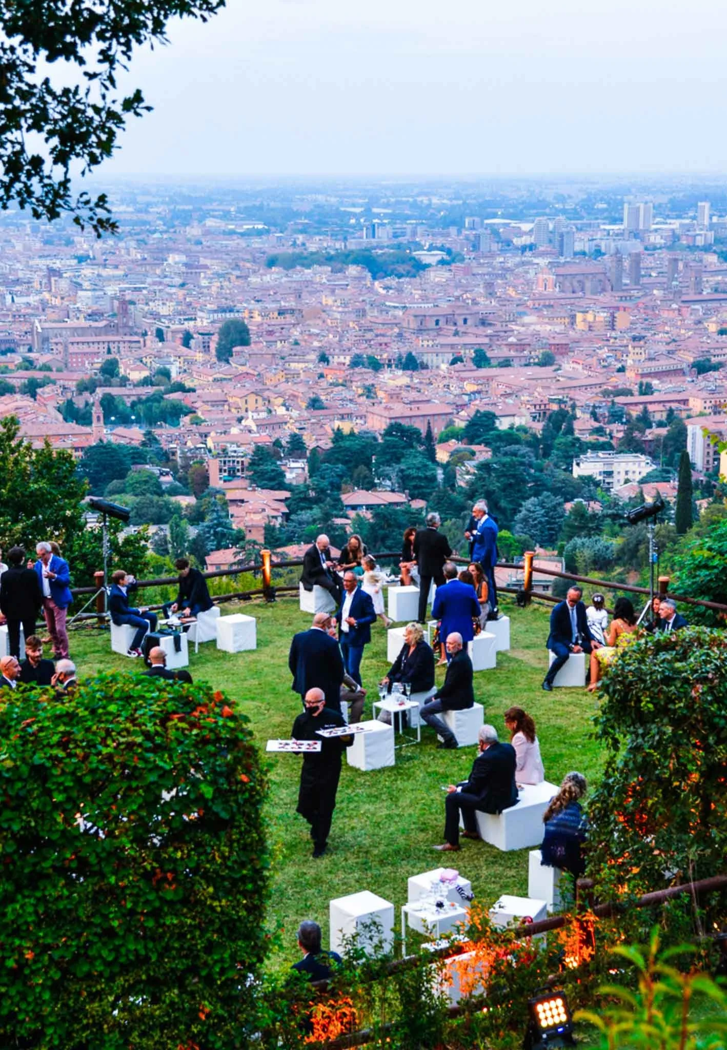 Una riunione o evento sociale con persone sedute e in piedi su un prato, con una vista panoramica sulla città di notte o sera.