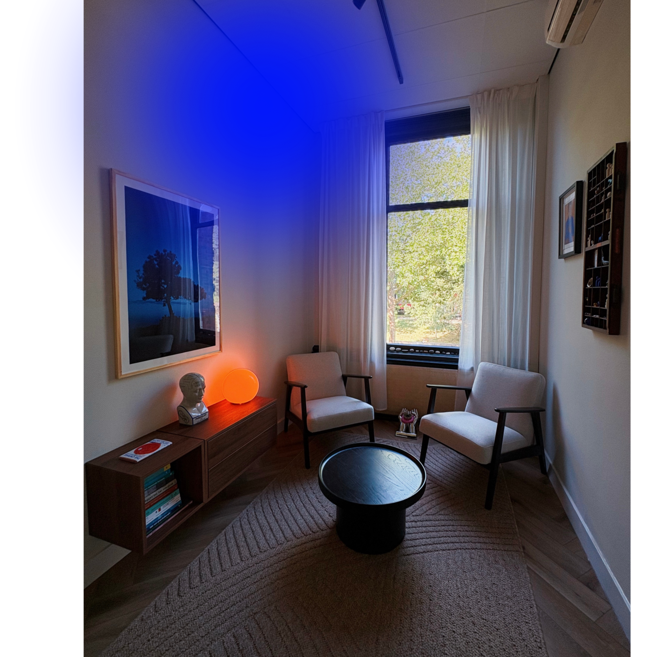 A cozy corner of a room with a window showing leafy trees outside. The wall has framed artwork and a shelf with books. There is a small table with a classical bust sculpture, an orange lamp, and a patterned rug.