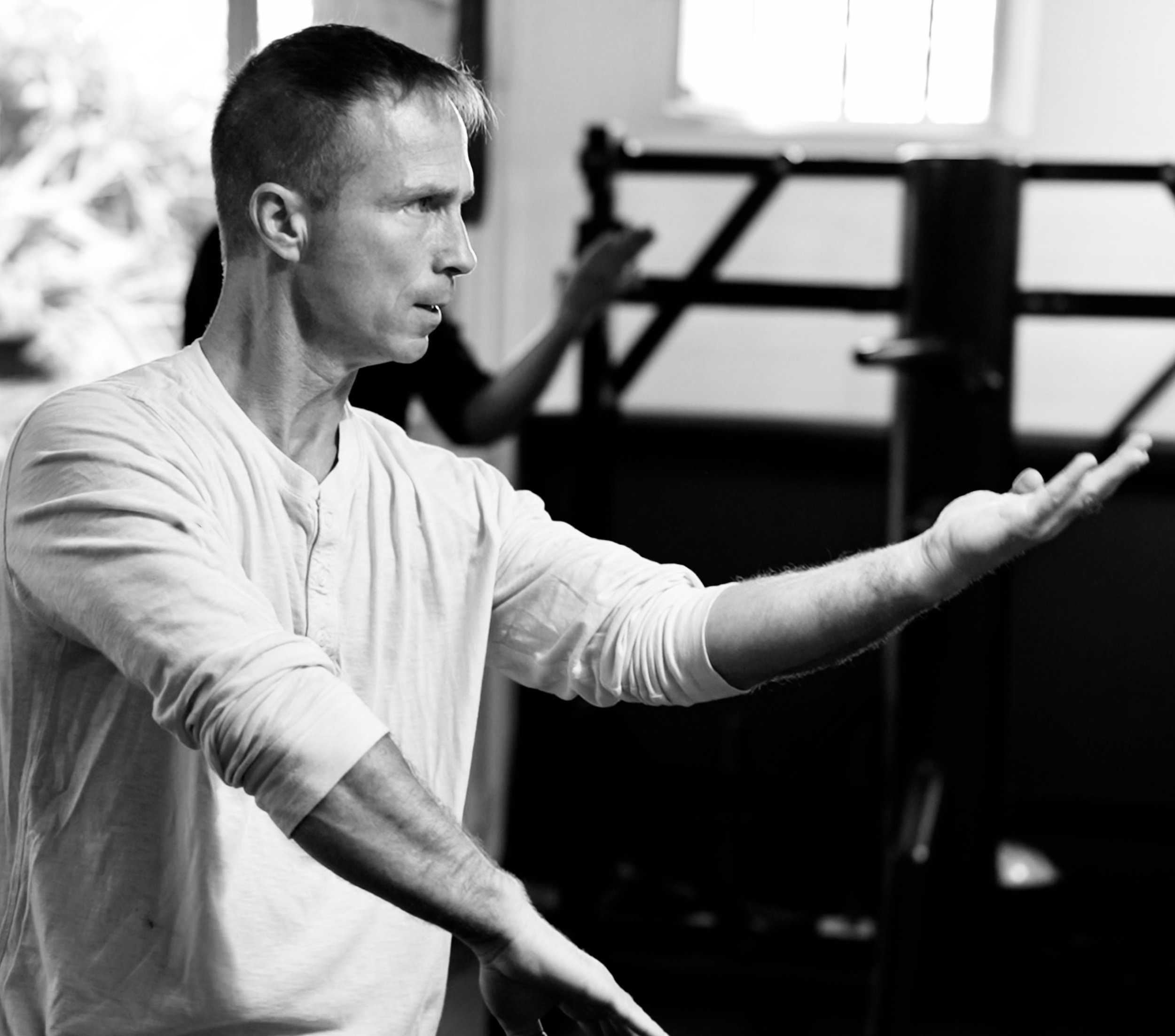 Un homme en train d'expliquer quelque chose, avec un bras tendu, dans une pièce avec des partitions musicales en arrière-plan.