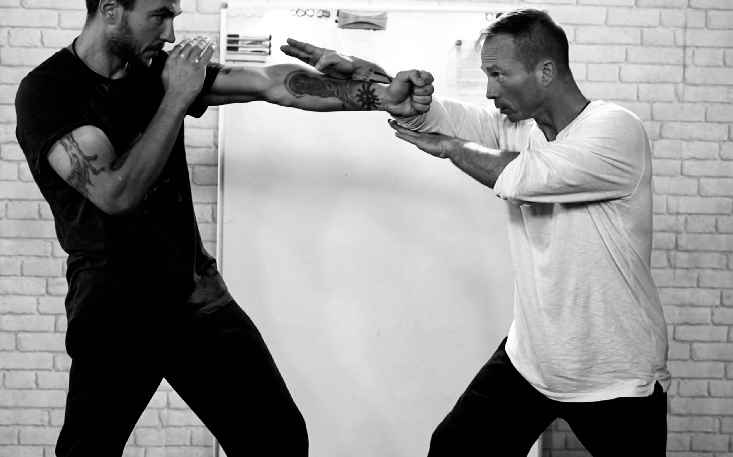 Deux hommes s'entraînent en combat de self-défense ou de boxe, l'un d'eux en position défensive, l'autre en train d'auditionner une frappe, dans une salle avec un mur en briques.