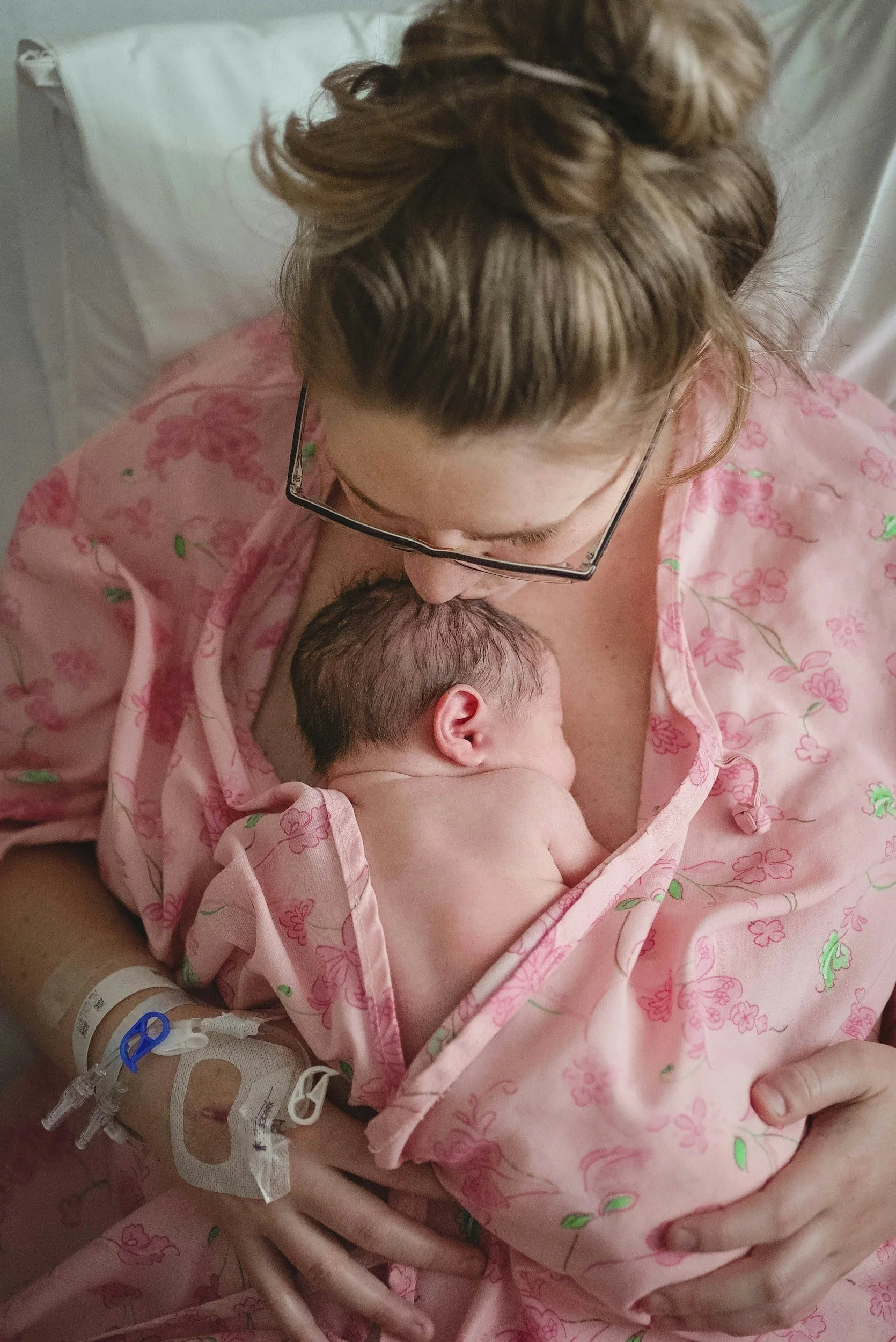 A woman with glasses holding a newborn baby against her chest in a hospital bed, both wrapped in a pink floral hospital gown, with the woman wearing an IV bandage on her arm.