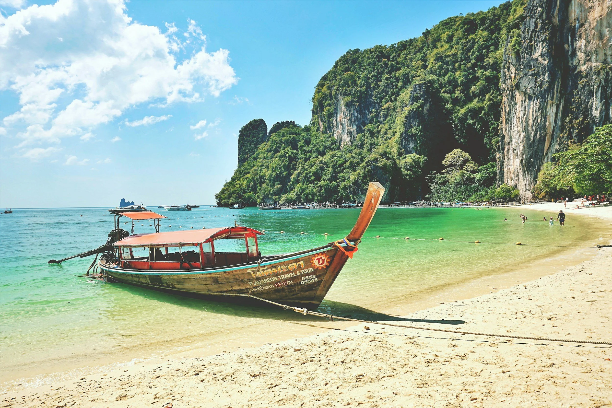 A traditional longtail boat on a sandy beach with turquoise water, lush green cliffs, and a partly cloudy sky.