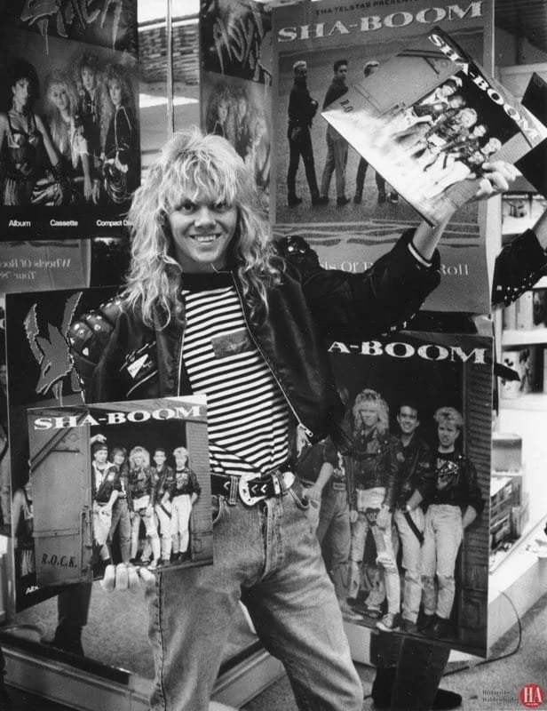 A woman with curly blonde hair smiling and holding a vinyl record, standing in front of posters advertising the band Sha-Boom with images of the band members.