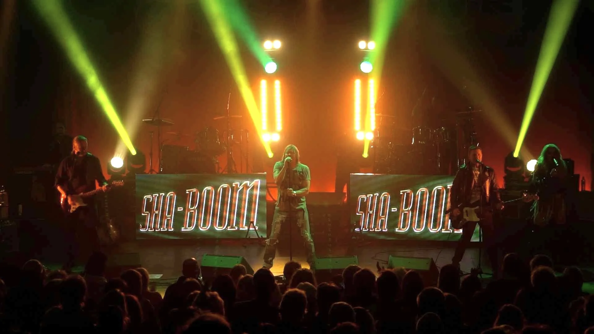 A music concert with a band performing on stage with neon signs that read 'SHA-Boo' and colorful stage lighting, including green and yellow spotlights, in front of an audience.