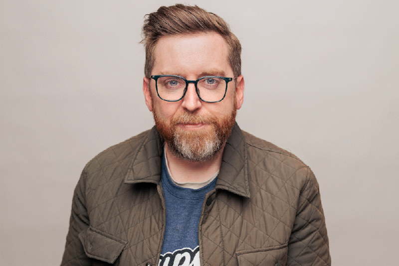 Rudy Harris with his glasses, a beard, and short hair, wearing a quilted jacket over a blue t-shirt, standing against a plain light gray background.