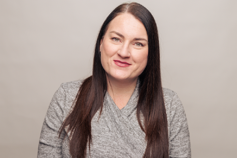 A woman with long brown hair wearing a gray sweater, smiling at the camera against a plain background.