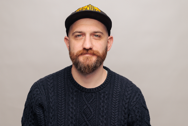 Ryan Neltner wearing a black knit sweater and a black cap with yellow text, standing against a plain light gray background.