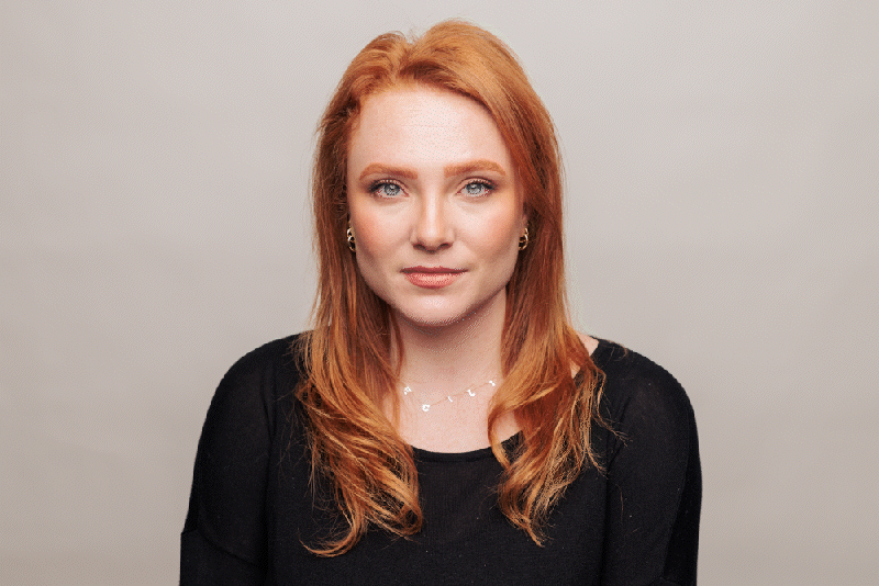 A woman with shoulder-length red hair and blue eyes, wearing a black top, sitting against a plain light gray background.