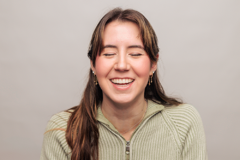 A young woman with long brown hair smiling and eyes closed, wearing a light green zip-up sweater and earrings, against a plain light gray background.