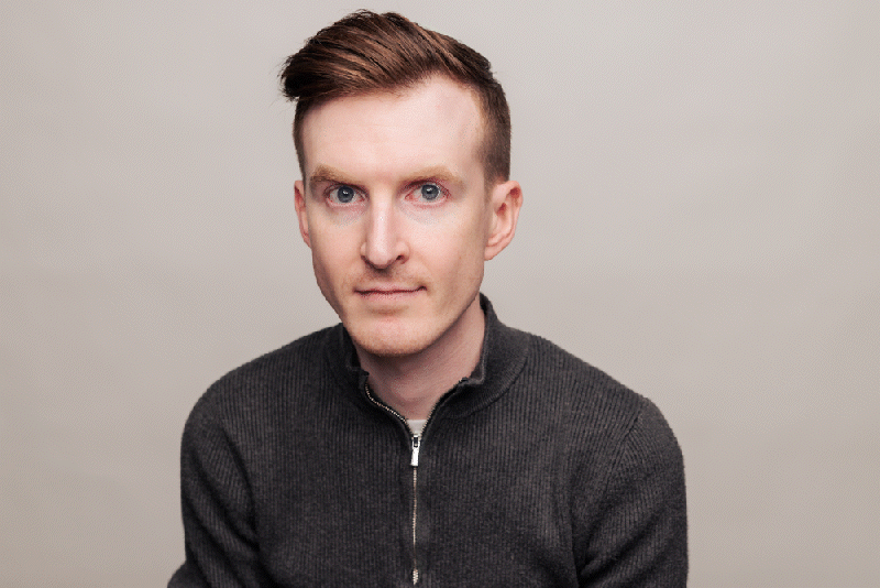 Portrait of a man with short brown hair, blue eyes, wearing a dark gray zip-up sweater, against a light gray background.