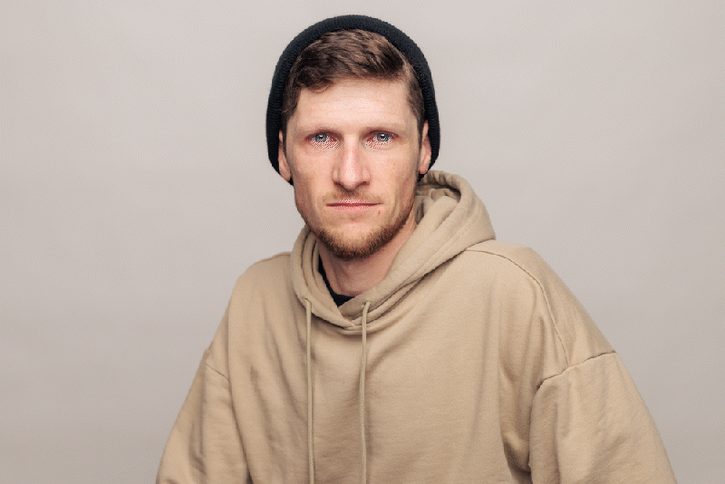 A young man with short brown hair, blue eyes, and a scruffy beard is wearing a black beanie and a beige hoodie, looking directly at the camera with a neutral expression against a plain gray background.
