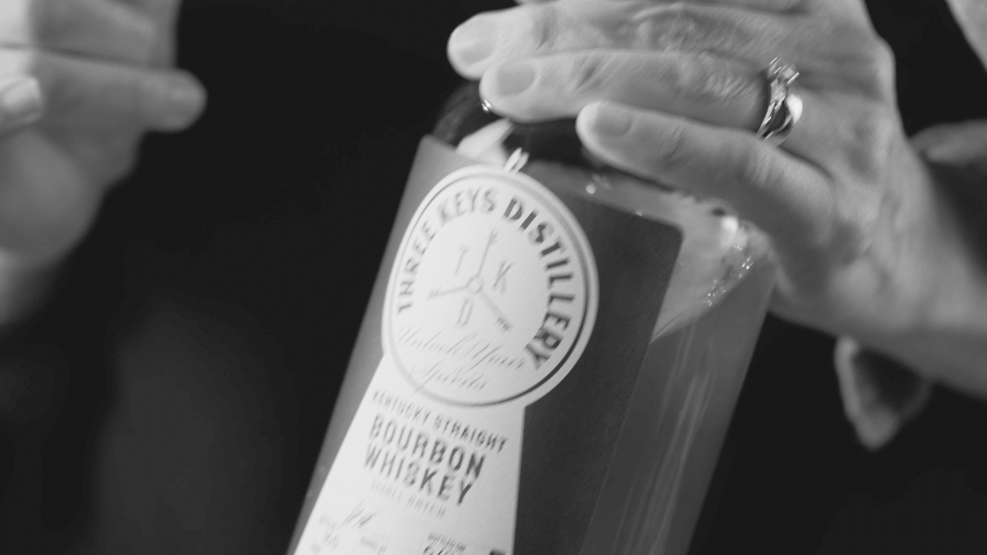 Close-up of a person's hand with a ring, opening a bottle of whiskey labeled 'Thirsty Keys Digital' Bourbon.