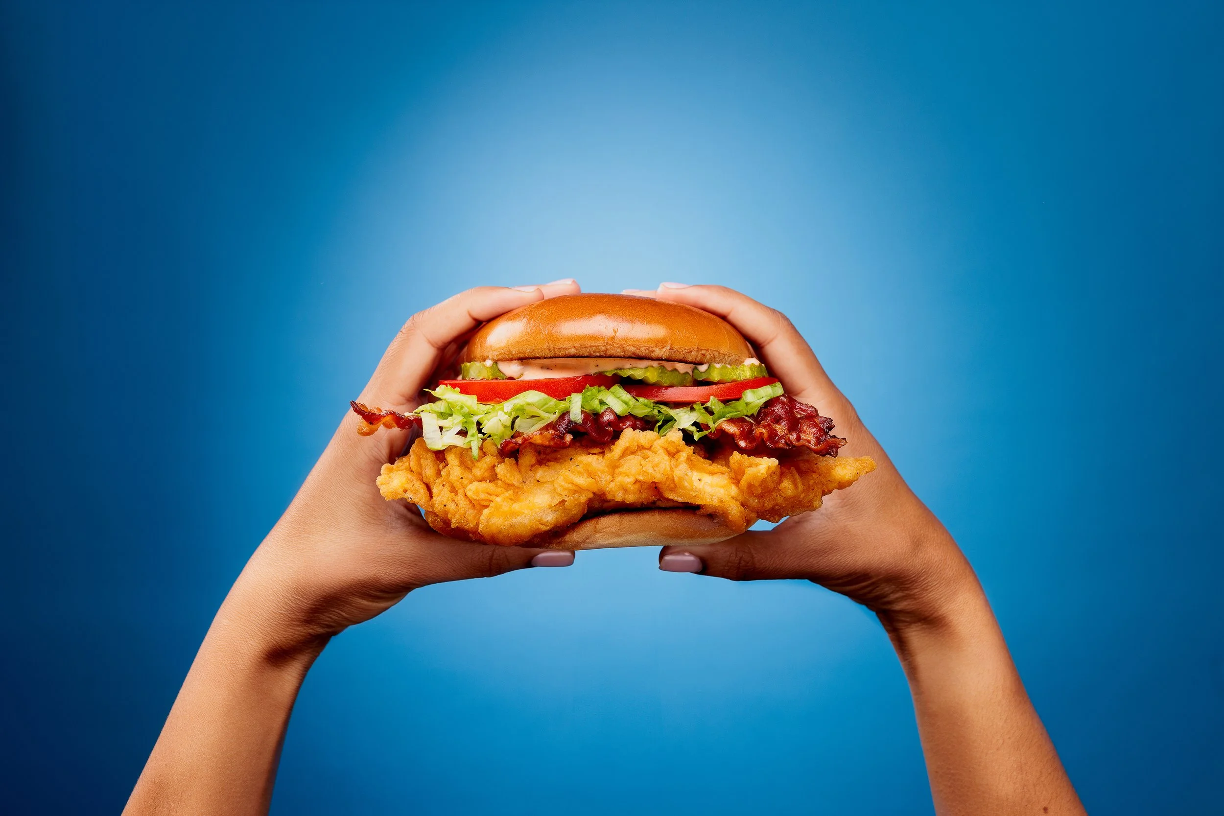 Person holding a fried chicken sandwich with lettuce, tomato, pickles, bacon, and a bun against a blue background.