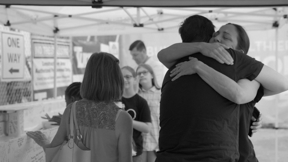 Two people hugging each other tightly at an outdoor event, surrounded by a group of onlookers.