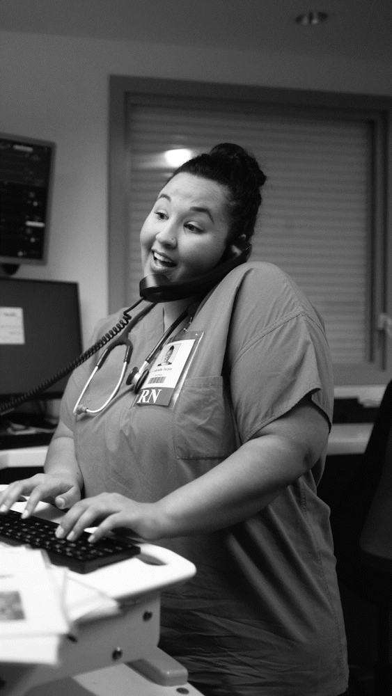 A female nurse in scrubs with a stethoscope around her neck, talking on the phone while working at a computer in a medical setting.
