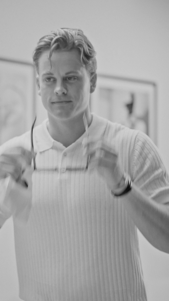 Black and white photo of a young man with styled hair, wearing a light-colored, long-sleeve, ribbed shirt, holding sunglasses in front of him, in an indoor setting with framed artwork on the wall.