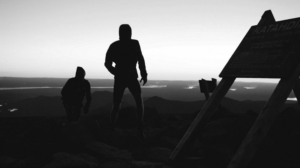 Silhouettes of two people hiking on a mountain during sunset, with a signboard in the foreground.
