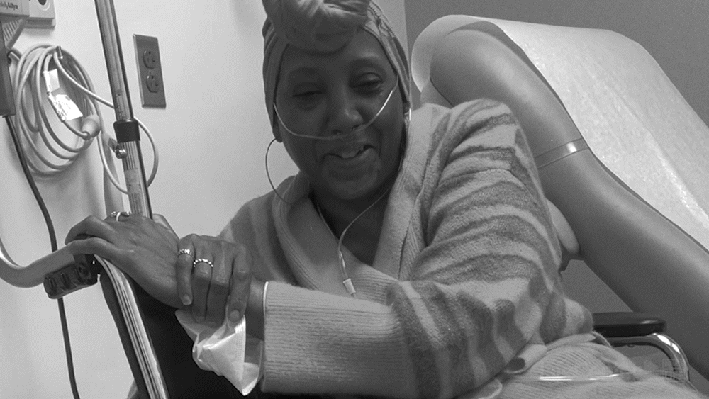 A woman in a hospital bed with an IV line and oxygen tube, smiling and holding the bed rail.