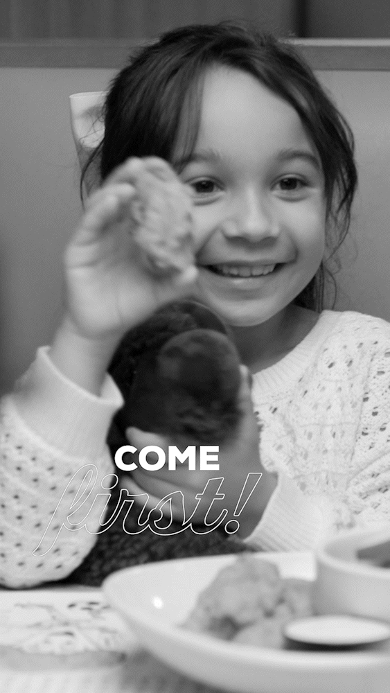 A young girl smiling and holding up a piece of food, with the words 'COME first!' overlaid on the image. She is wearing a patterned sweater and sitting at a table with food in front of her.