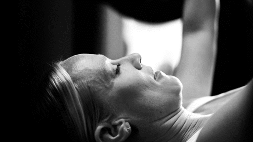 A woman lying on her back with her eyes closed while a person behind her touches her forehead.