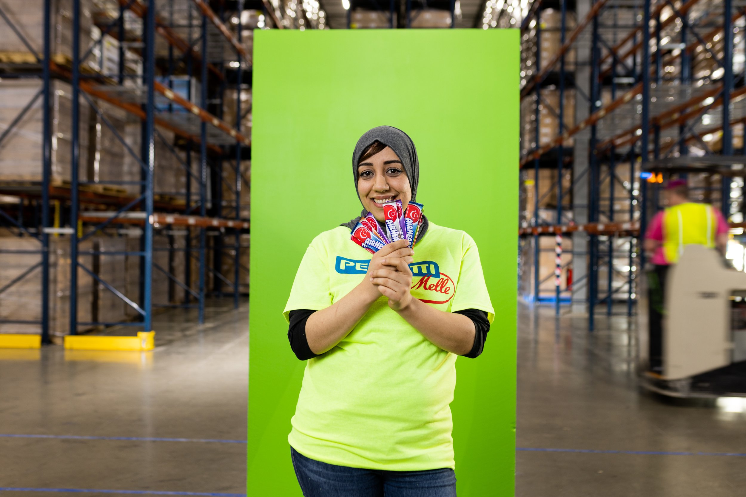 A smiling woman wearing a gray hijab and a yellow shirt, holding several Aero chocolate bars, standing in front of a green backdrop inside a warehouse.