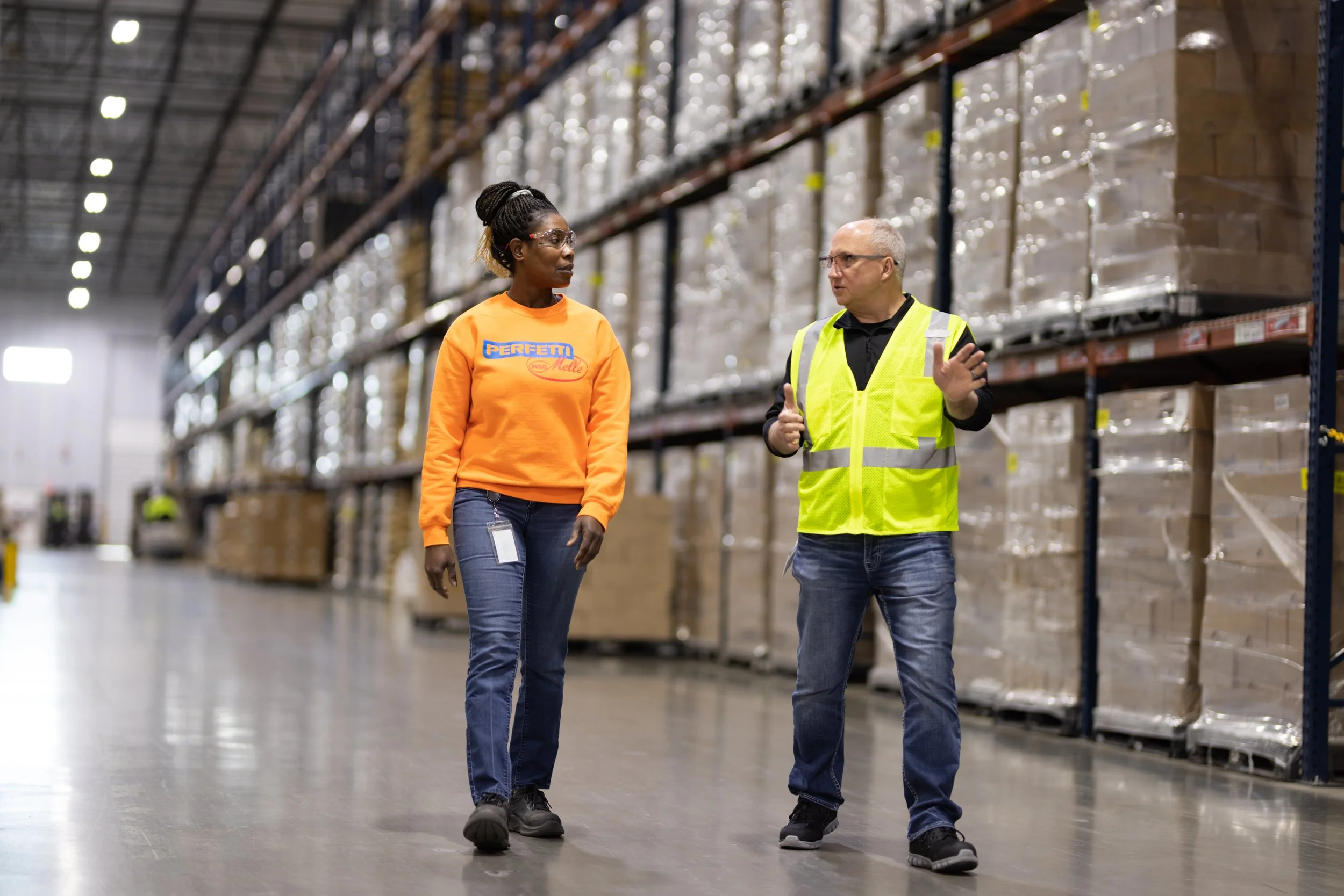 Two warehouse workers, a woman and a man, having a conversation while walking through an aisle filled with shelves stocked with boxes and pallets in a large industrial warehouse. The man is wearing a high-visibility vest and gesturing with his hand, 