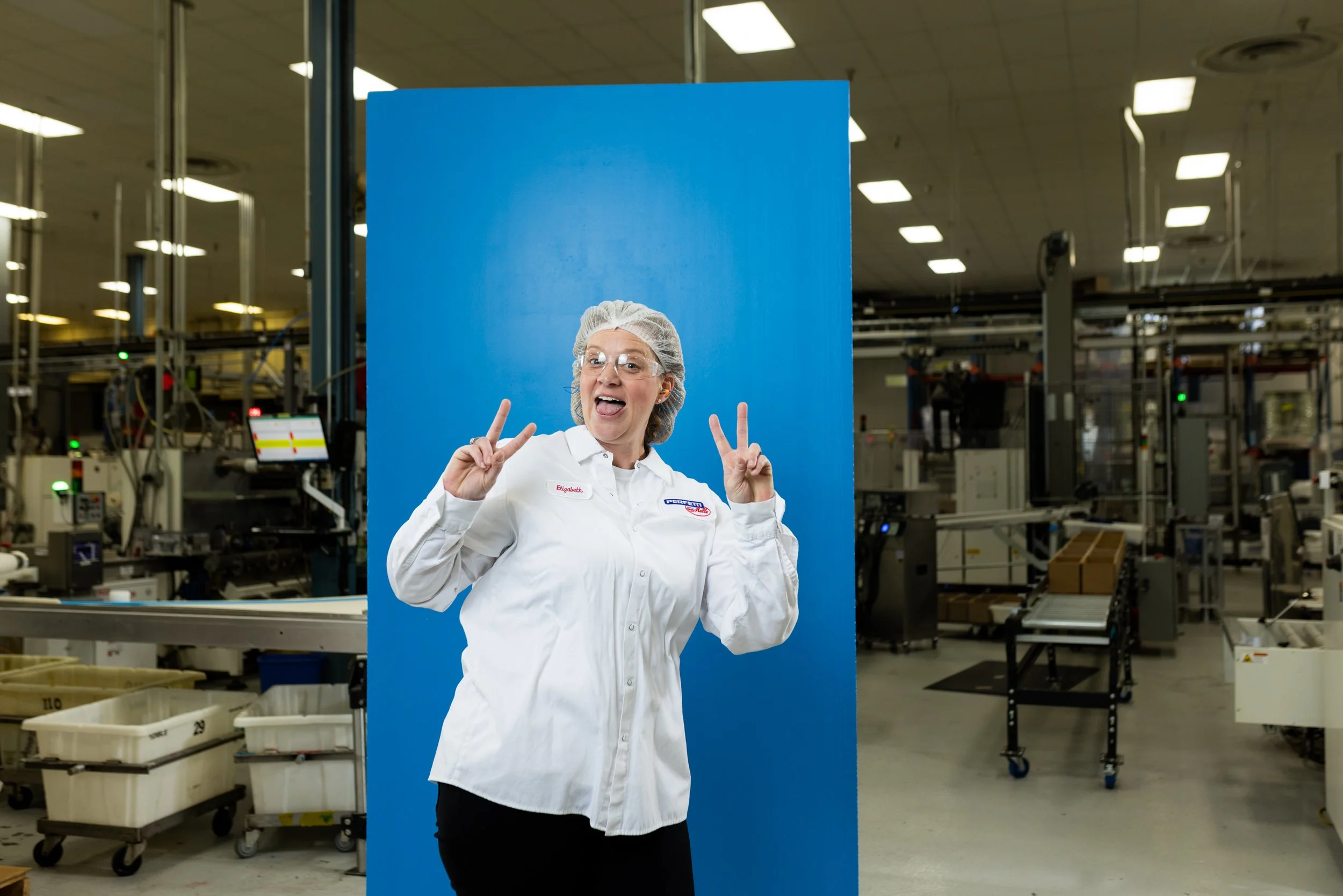 A woman in a white work uniform and hairnet making peace signs with both hands inside an industrial manufacturing facility.