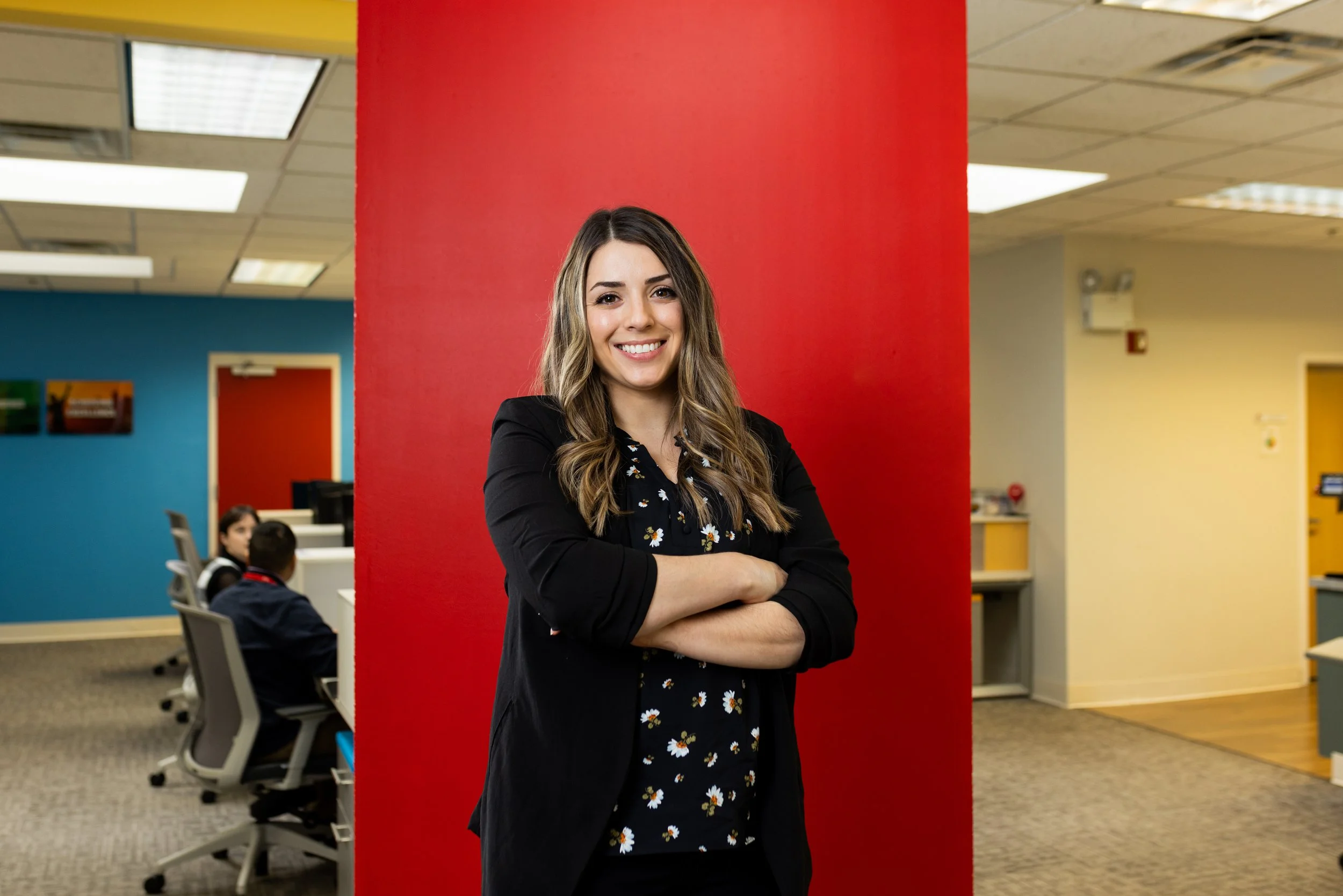 A woman with long wavy brown hair, wearing a black blazer and a black blouse with white daisy print, standing in front of a red wall inside an office, smiling with arms crossed.