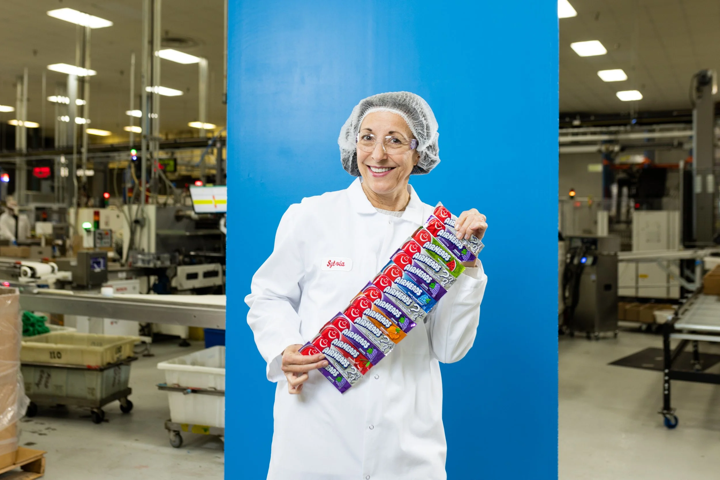 A woman in a white lab coat and hair net holding multiple packs of Airheads candy in a factory setting.