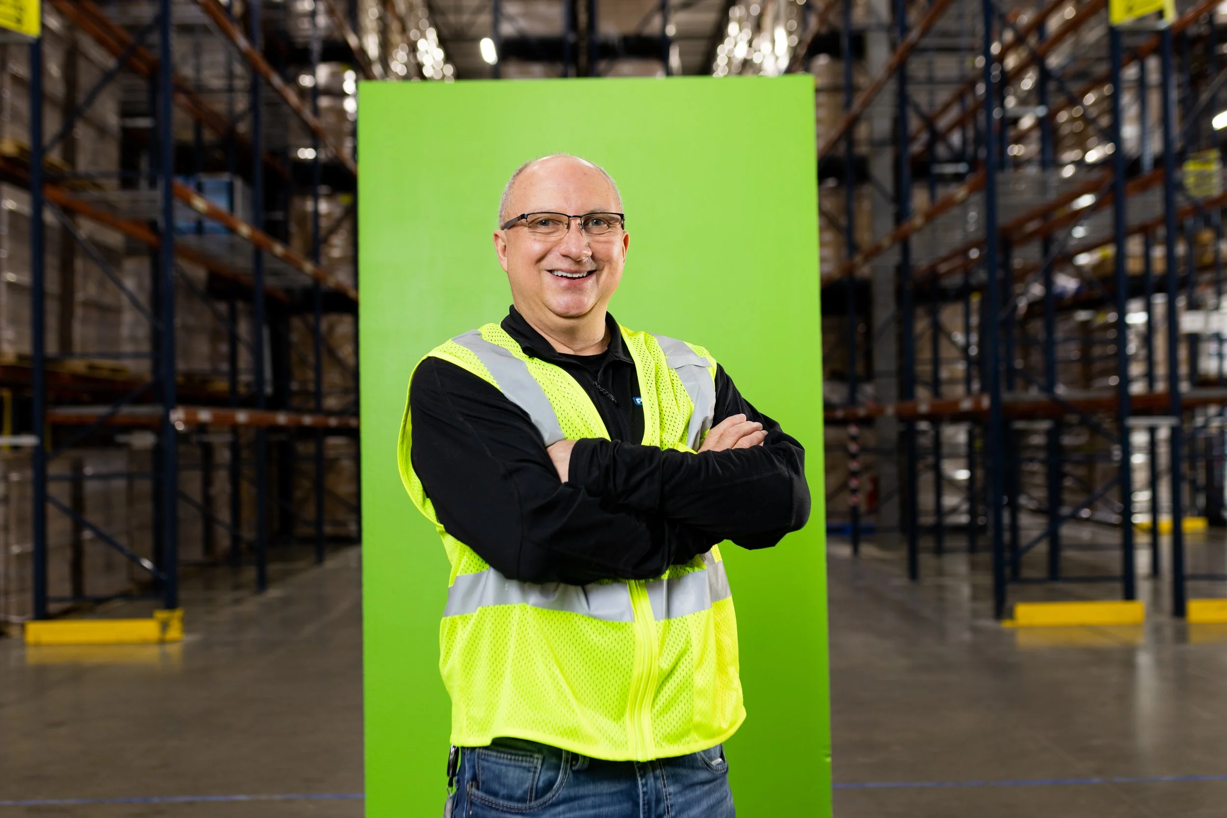 A man smiling with arms crossed, wearing a black shirt and a yellow safety vest, standing in a warehouse with shelving units in the background and a green screen behind him.