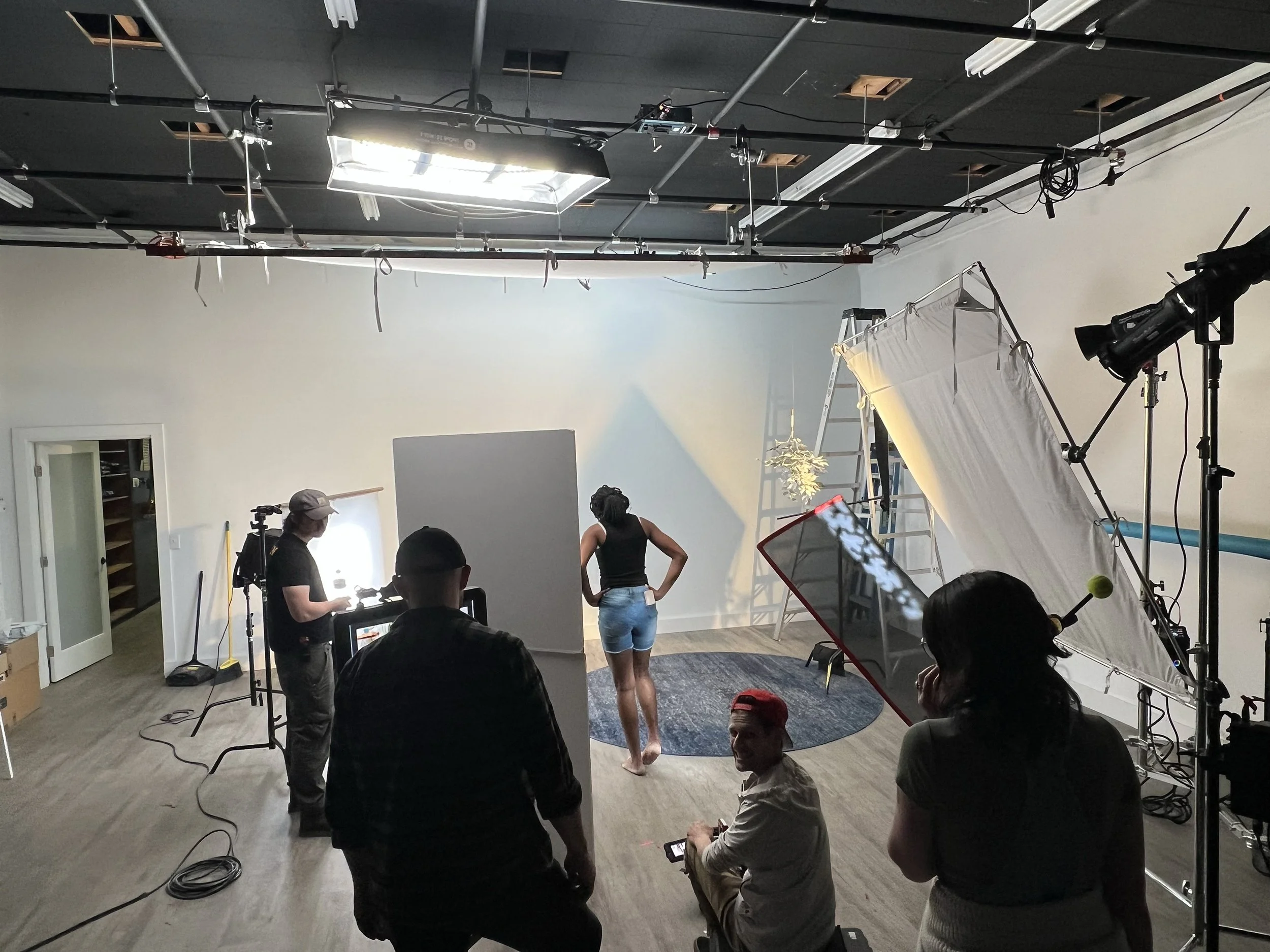 Behind the scenes of a photo shoot in a studio with a woman posing, professional lighting, a white backdrop, and crew members preparing equipment.