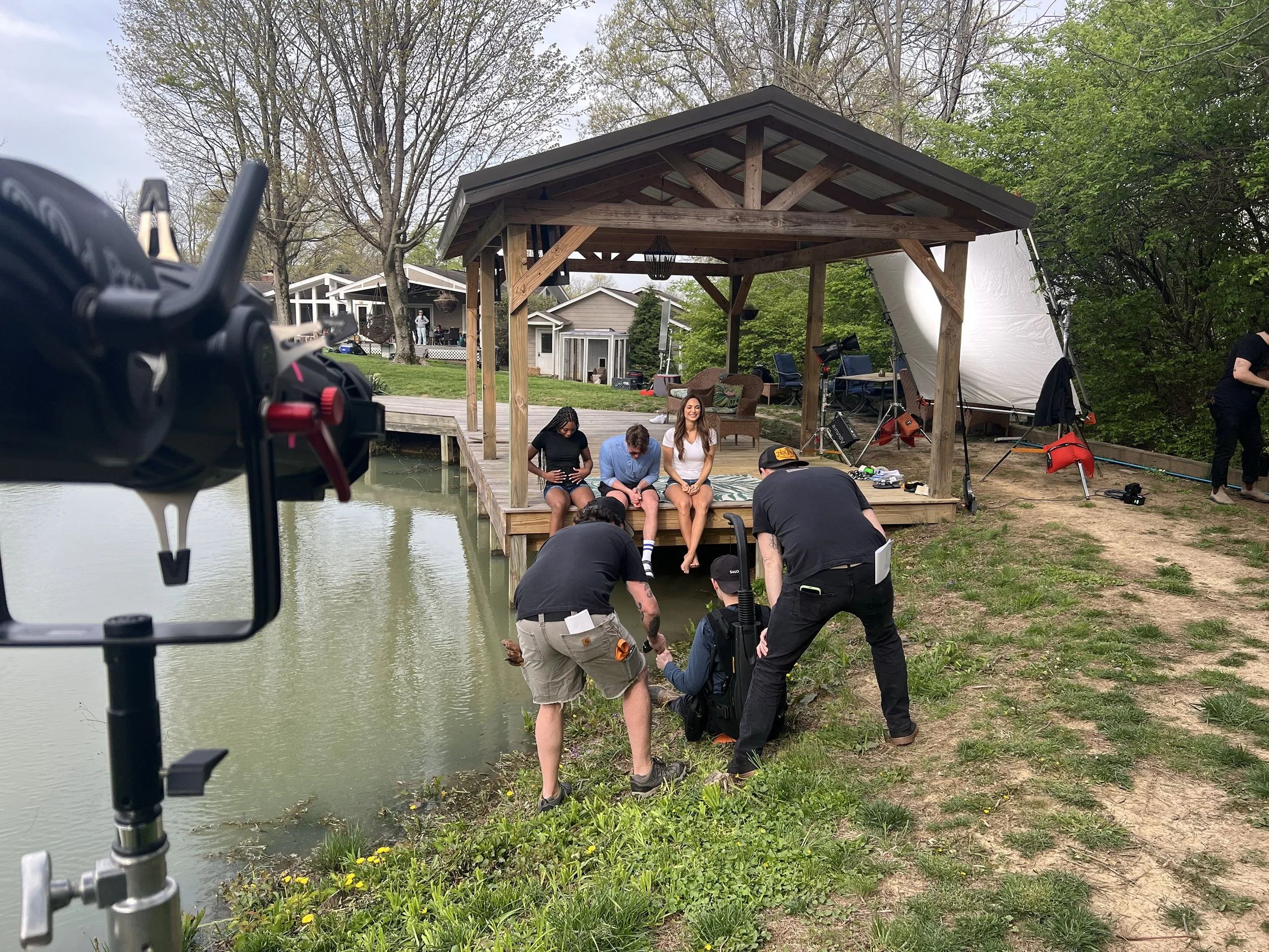A behind-the-scenes photo of a film or photo shoot near a small lake, with a crew capturing images of three women sitting on a wooden dock under a gazebo. The crew is working on the grass by the water's edge, with camera equipment and crew members vi