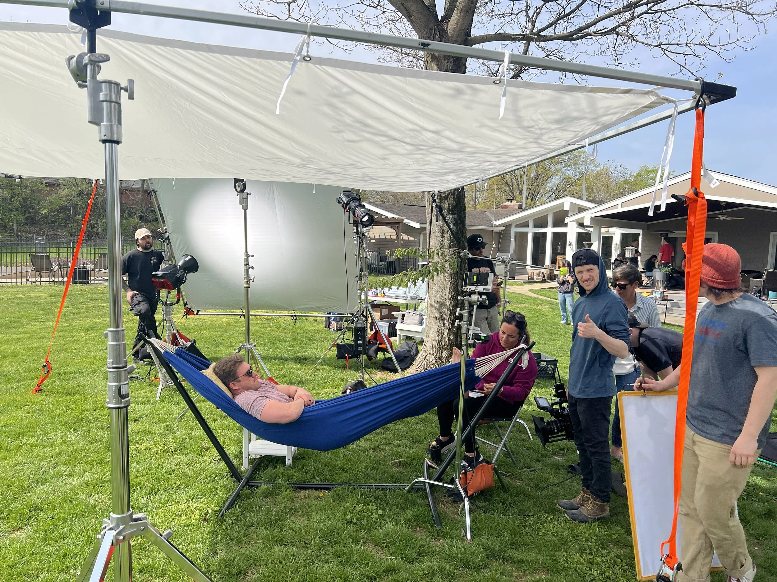 A film crew setting up for a shoot outdoors, with a woman lying in a hammock surrounded by equipment, crew members, and a large reflector. A house and other crew members are visible in the background.