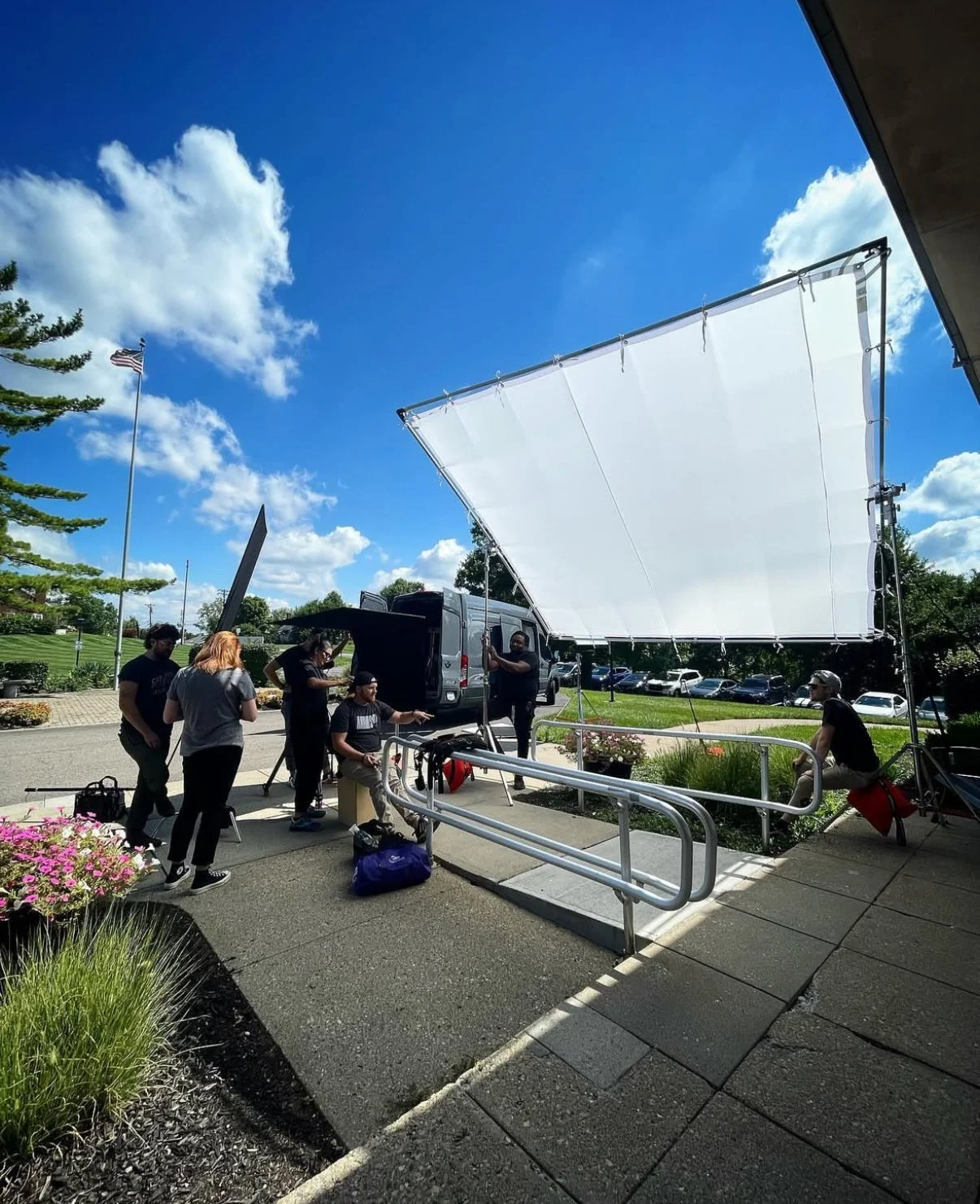 A film crew setting up outdoor filming equipment on a bright, sunny day with a blue sky filled with clouds. Crew members are working near a large white diffuser, with some standing and others sitting or handling equipment, surrounded by parked cars a
