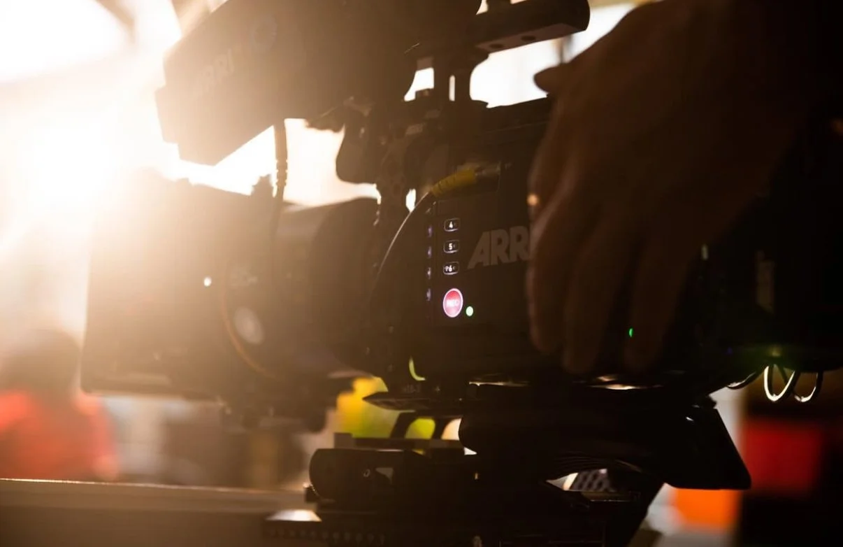 Close-up of a professional video camera mounted on a tripod with a person's hand adjusting settings, backlit by bright sunlight.